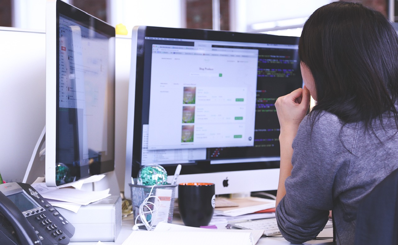 A woman working on a computer with multiple screens displaying code and a web application, representing IT project management and budgeting challenges.